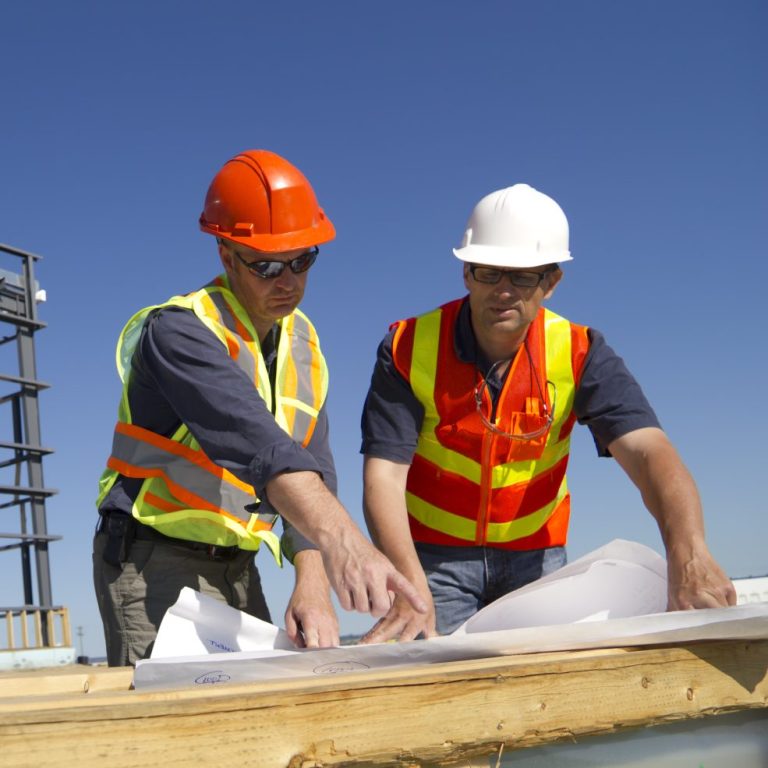 Zwei Bauarbeiter mit Helmen und Warnwesten, die Pläne auf einer Baustelle prüfen.