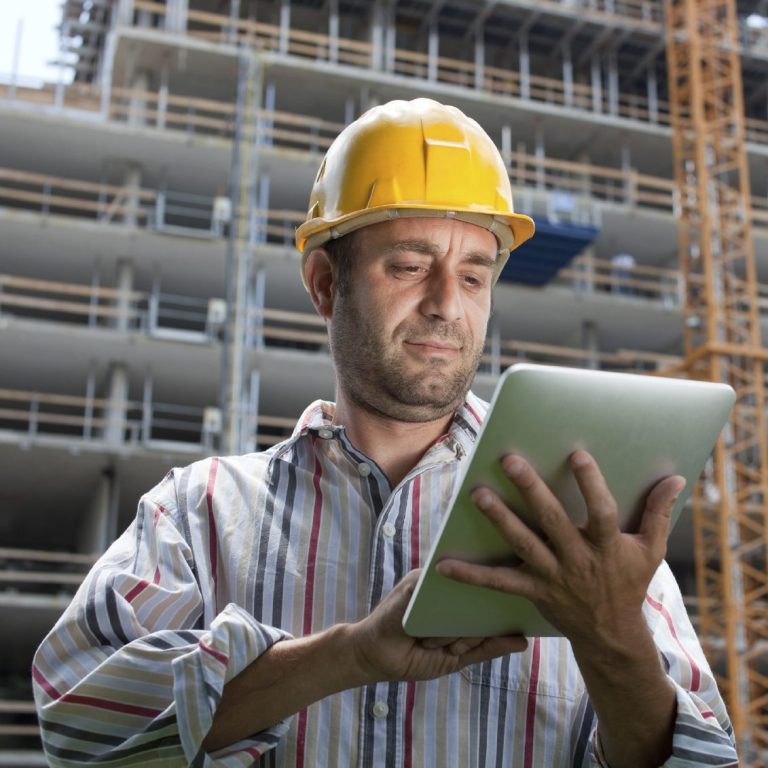 Mann in Helm nutzt ein Tablet auf einer Baustelle mit Gebäudestruktur im Hintergrund.